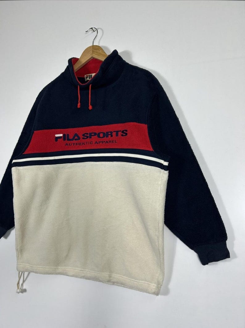 May include: A navy blue, red, and cream-colored FILA SPORTS fleece pullover. The pullover features a high collar, long sleeves, and the FILA SPORTS logo in red across the chest. The bottom half is cream-colored, with a white stripe separating it from the red band.