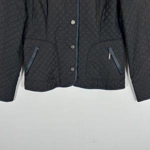 May include: Black quilted jacket with a diamond pattern. Features silver buttons, two front pockets with navy blue trim, and a navy blue trim along the collar. A classic, stylish outerwear piece.