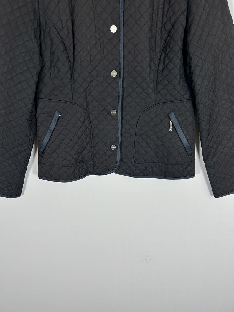 May include: Black quilted jacket with a diamond pattern. Features silver buttons, two front pockets with navy blue trim, and a navy blue trim along the collar. A classic, stylish outerwear piece.