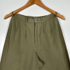 Puede incluir: Pantalones verde oliva con cierre de bot&oacute;n y cremallera, colgados de una percha de madera. Los pantalones tienen pliegues en la cintura y un corte relajado. La tela parece ser un algod&oacute;n ligero o mezcla.