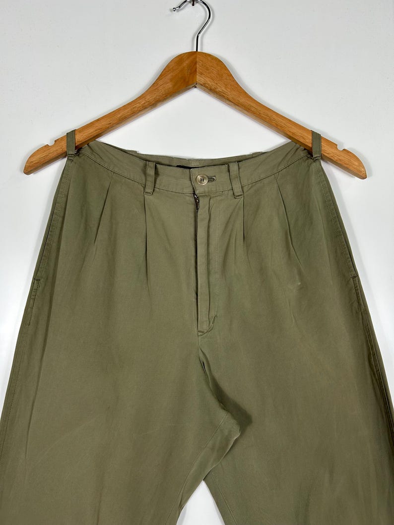 Puede incluir: Pantalones verde oliva con cierre de bot&oacute;n y cremallera, colgados de una percha de madera. Los pantalones tienen pliegues en la cintura y un corte relajado. La tela parece ser un algod&oacute;n ligero o mezcla.