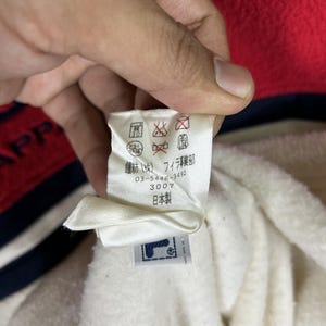 May include: A close-up of a white tag with Japanese text and a FILA logo. The tag is attached to a white fabric, with red and navy blue fabric visible in the background. The tag includes care symbols.