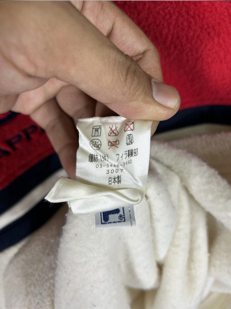 May include: A close-up of a white tag with Japanese text and a FILA logo. The tag is attached to a white fabric, with red and navy blue fabric visible in the background. The tag includes care symbols.