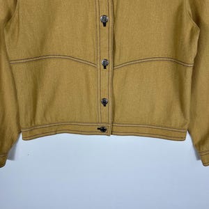 Peut inclure: Une veste jaune moutarde avec des surpiq&ucirc;res contrastantes marron fonc&eacute;, une fermeture boutonn&eacute;e avec des boutons argent&eacute;s m&eacute;talliques et des coutures horizontales sur la poitrine et le torse. La veste a des manches longues avec poignets boutonn&eacute;s.