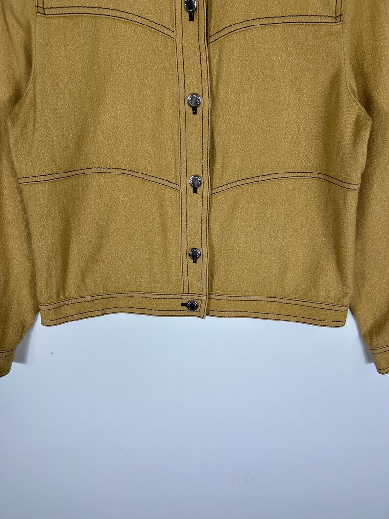 Peut inclure: Une veste jaune moutarde avec des surpiq&ucirc;res contrastantes marron fonc&eacute;, une fermeture boutonn&eacute;e avec des boutons argent&eacute;s m&eacute;talliques et des coutures horizontales sur la poitrine et le torse. La veste a des manches longues avec poignets boutonn&eacute;s.