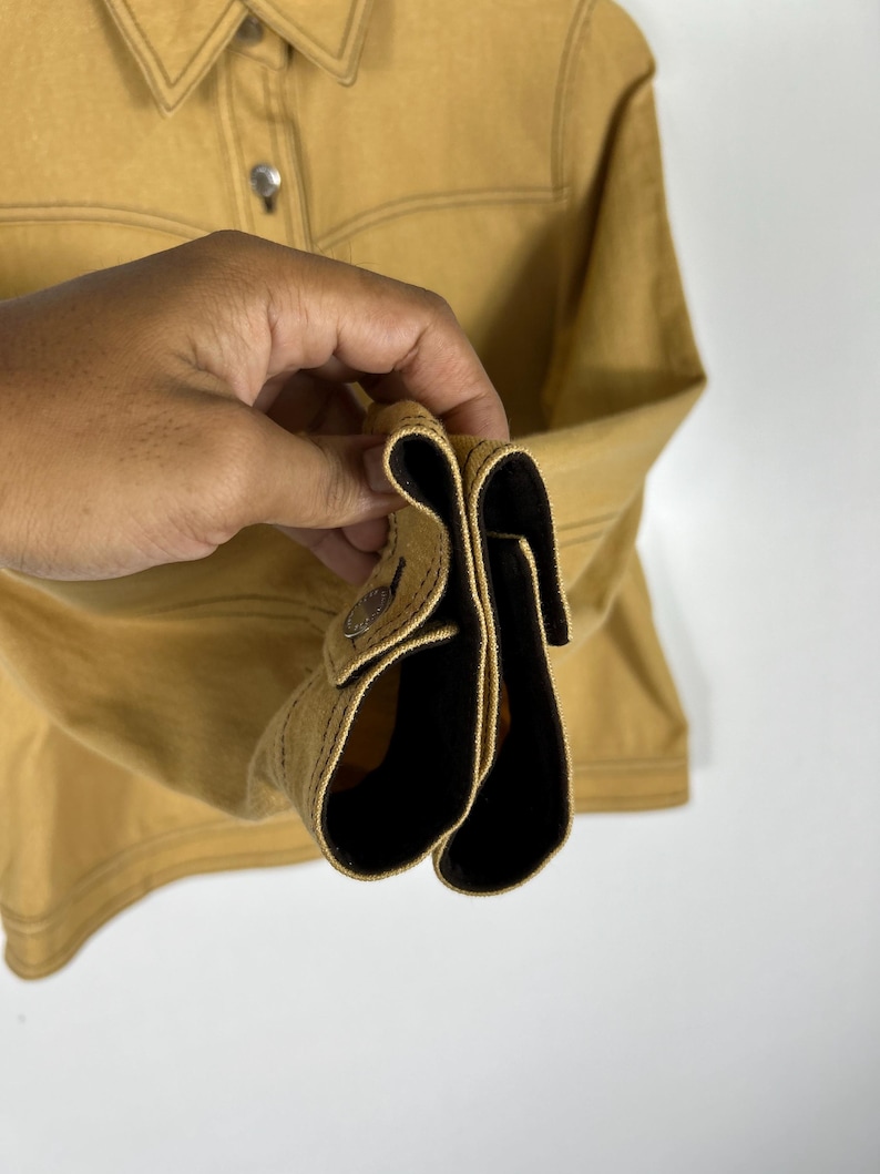Peut inclure: Une chemise &agrave; manches longues jaune moutarde dot&eacute;e d'un col pointu, d'une patte de boutonnage avant avec des boutons m&eacute;talliques et de boutons m&eacute;talliques assortis aux poignets. Le v&ecirc;tement pr&eacute;sente des surpiq&ucirc;res visibles le long des coutures.