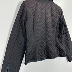 May include: A black quilted jacket with a diamond pattern. The jacket has a collar and long sleeves. The cuffs and collar have a dark trim. The jacket is hanging on a wooden hanger against a white background.