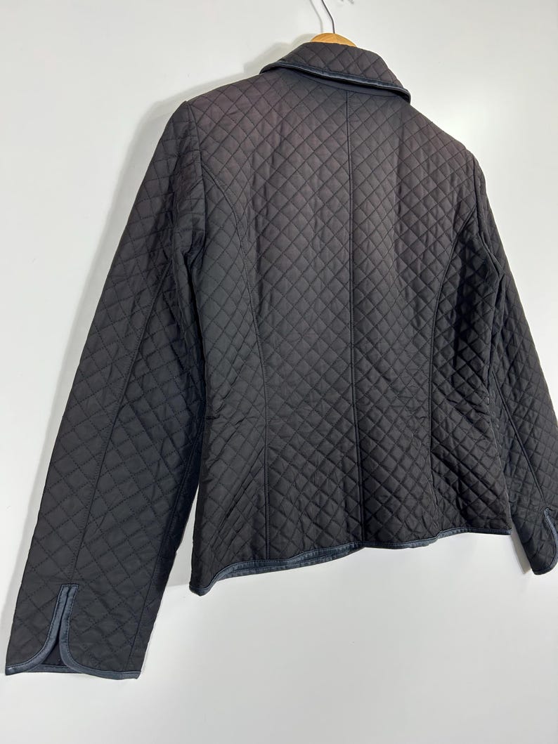 May include: A black quilted jacket with a diamond pattern. The jacket has a collar and long sleeves. The cuffs and collar have a dark trim. The jacket is hanging on a wooden hanger against a white background.
