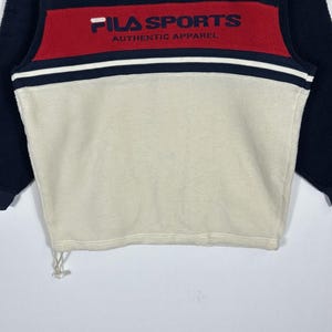 May include: A navy blue, red, and cream-colored fleece pullover. The top features a red band across the chest with "FILA SPORTS AUTHENTIC APPAREL" printed in white. The pullover has a drawstring at the bottom and navy blue sleeves.