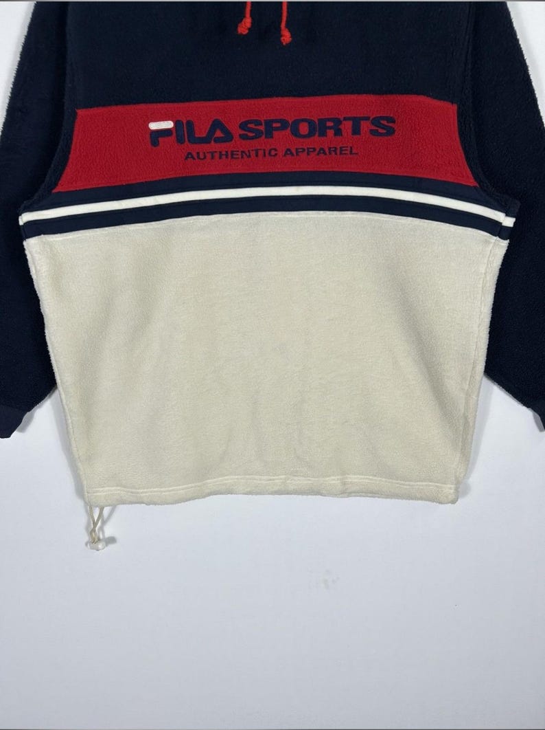 May include: A navy blue, red, and cream-colored fleece pullover. The top features a red band across the chest with "FILA SPORTS AUTHENTIC APPAREL" printed in white. The pullover has a drawstring at the bottom and navy blue sleeves.