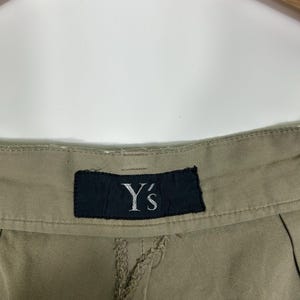 Puede incluir: Primer plano de unos pantalones color caqui con una costura visible y una etiqueta negra. La etiqueta muestra el texto blanco "Y's". Los pantalones cuelgan de una percha de madera sobre un fondo blanco.