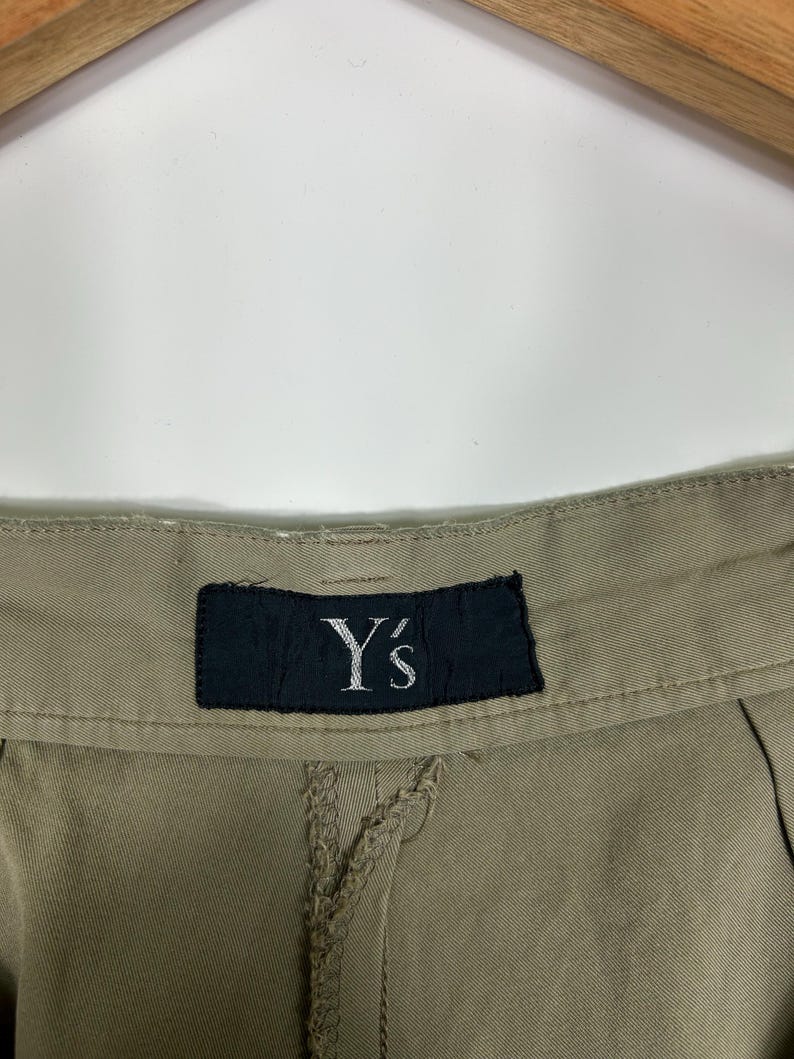 Puede incluir: Primer plano de unos pantalones color caqui con una costura visible y una etiqueta negra. La etiqueta muestra el texto blanco "Y's". Los pantalones cuelgan de una percha de madera sobre un fondo blanco.