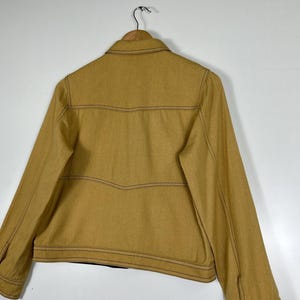 Peut inclure: Une veste jaune moutarde dot&eacute;e d'un col classique, de manches longues avec poignets boutonn&eacute;s et de surpiq&ucirc;res contrastantes fonc&eacute;es visibles sur tout le panneau arri&egrave;re. Le v&ecirc;tement est pr&eacute;sent&eacute; sur un cintre en bois sur fond blanc.