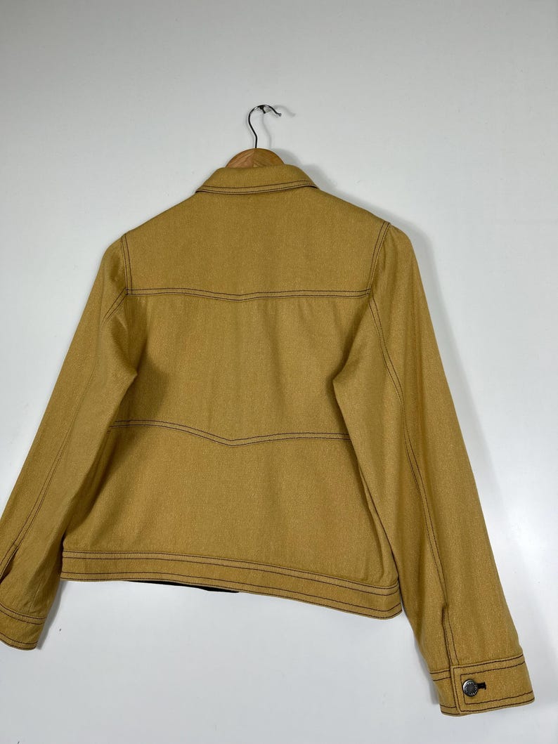 Peut inclure: Une veste jaune moutarde dot&eacute;e d'un col classique, de manches longues avec poignets boutonn&eacute;s et de surpiq&ucirc;res contrastantes fonc&eacute;es visibles sur tout le panneau arri&egrave;re. Le v&ecirc;tement est pr&eacute;sent&eacute; sur un cintre en bois sur fond blanc.