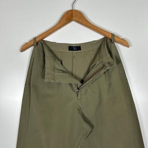 Puede incluir: Pantalones verde oliva colgados de una percha de madera. Los pantalones tienen cremallera y cierre de bot&oacute;n. La marca "Y's" es visible en la cintura. Los pantalones est&aacute;n hechos de una tela duradera y tienen un corte relajado.