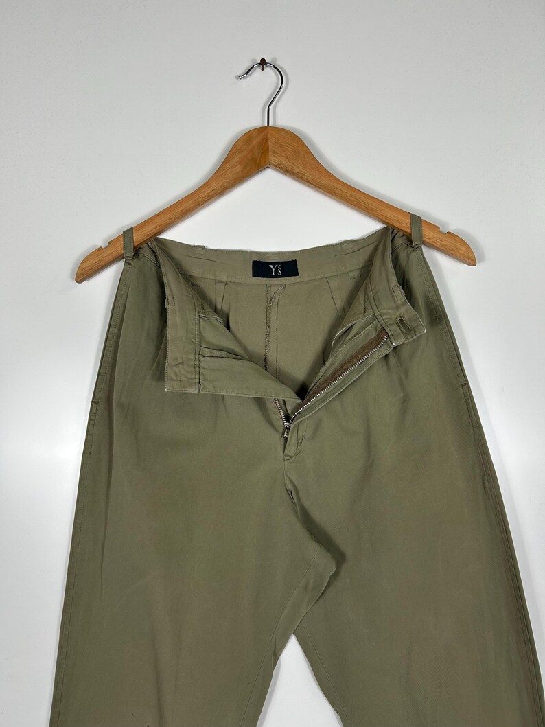 Puede incluir: Pantalones verde oliva colgados de una percha de madera. Los pantalones tienen cremallera y cierre de bot&oacute;n. La marca "Y's" es visible en la cintura. Los pantalones est&aacute;n hechos de una tela duradera y tienen un corte relajado.