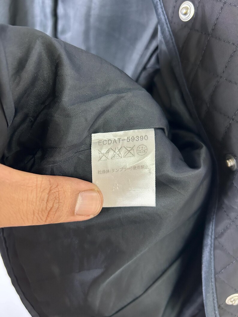 May include: Close-up of a black quilted jacket interior. The inside lining is black and smooth. A white tag with the text "ECDAT-59390" and care symbols is visible. The jacket has silver snap closures.