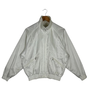May include: A white, vintage-style track jacket with a high collar and a full-length zipper. The jacket has two front pockets and ribbed cuffs and hem. The fabric has a subtle sheen.