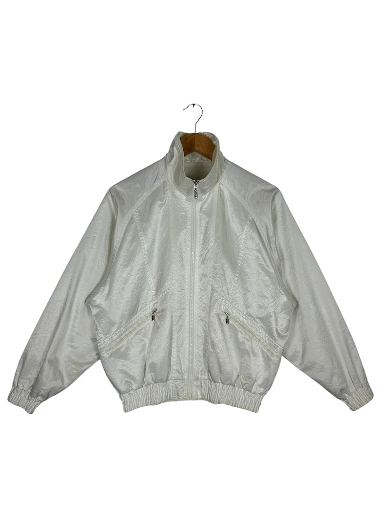 May include: A white, vintage-style track jacket with a high collar and a full-length zipper. The jacket has two front pockets and ribbed cuffs and hem. The fabric has a subtle sheen.