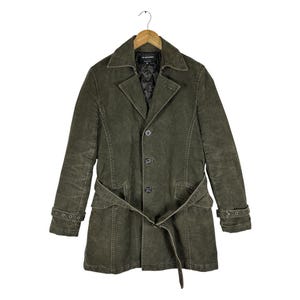 Op de afbeelding: Een olijfgroene corduroy jas met een reverskraag, een sluiting met drie knopen aan de voorzijde en een bijpassende strikceintuur in de taille. De mouwen hebben verstelbare gespriemen bij de manchetten en de binnenzijde is zwart doorgestikt.