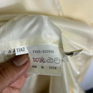 May include: A white garment care label featuring the text 11A2 and ECUS-100990, along with laundry symbols indicating no washing, no bleaching, and dry cleaning instructions. The label also specifies 100% wool content in Japanese text.