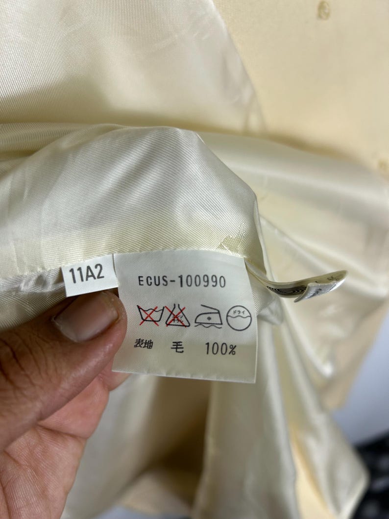 May include: A white garment care label featuring the text 11A2 and ECUS-100990, along with laundry symbols indicating no washing, no bleaching, and dry cleaning instructions. The label also specifies 100% wool content in Japanese text.