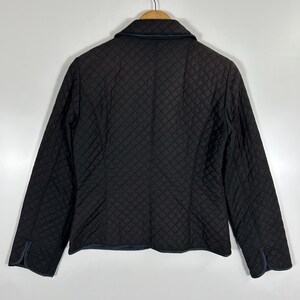 May include: A black quilted jacket with a classic collar and long sleeves. The jacket features a diamond pattern throughout and is hanging on a wooden hanger. The cuffs and collar have a dark blue trim.