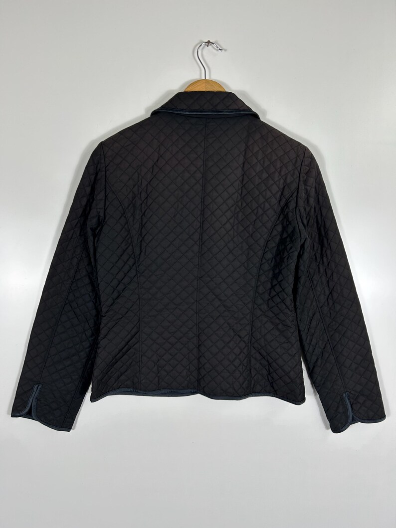 May include: A black quilted jacket with a classic collar and long sleeves. The jacket features a diamond pattern throughout and is hanging on a wooden hanger. The cuffs and collar have a dark blue trim.