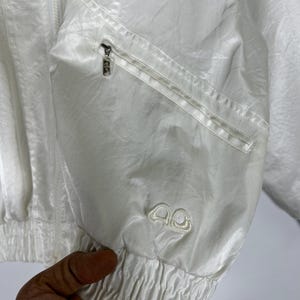 May include: A white, lightweight jacket with a zipper closure and a small zippered pocket. The jacket has a ribbed cuff and a small embroidered logo on the sleeve. The fabric has a subtle sheen.