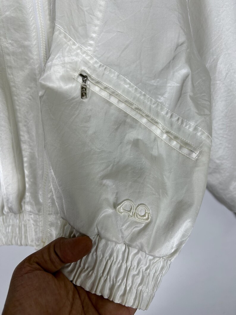 May include: A white, lightweight jacket with a zipper closure and a small zippered pocket. The jacket has a ribbed cuff and a small embroidered logo on the sleeve. The fabric has a subtle sheen.