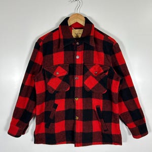 May include: A red and black checked wool jacket with a collar, two chest pockets, and two side pockets. The jacket has gold-coloured buttons and is hanging on a wooden hanger.