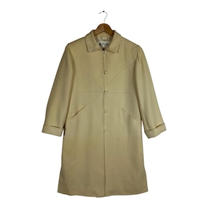 May include: A cream-coloured coat featuring a classic collar, long sleeves with folded cuffs, and a front button closure. The garment is accented with decorative curved seam detailing across the chest and torso, and it hangs on a wooden hanger.