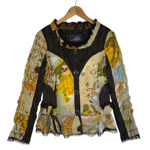 Op de afbeelding: Een vintage jasje met een uniek patchwork-ontwerp. De jas heeft een mix van bloemen- en abstracte patronen in geel, bruin en blauw. Het heeft een gerimpelde kraag en zoom, met zwarte accenten en knoopsluitingen. De jas hangt aan een houten kleerhanger.