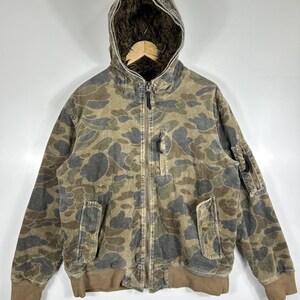 May include: A camouflage-patterned jacket with a hood and a zip-up front. The jacket features a brown ribbed hem and cuffs, two front pockets, and a small pocket on the left sleeve. The hood has a faux fur lining.