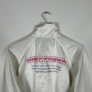 May include: White windbreaker jacket with a high collar, hanging on a metal hanger. The back of the jacket features the brand name "Courr&egrave;ges" in pink letters, along with additional text in a smaller font. The jacket has a shiny, textured appearance.