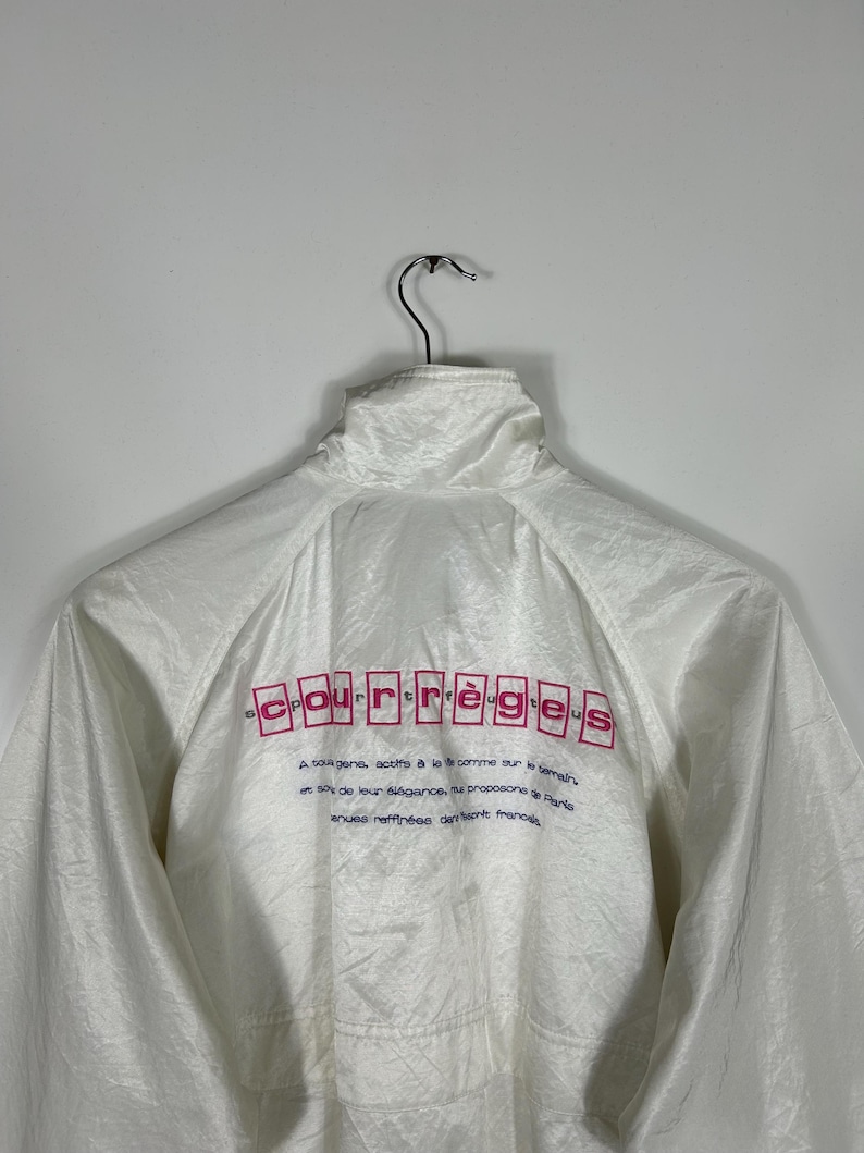 May include: White windbreaker jacket with a high collar, hanging on a metal hanger. The back of the jacket features the brand name "Courr&egrave;ges" in pink letters, along with additional text in a smaller font. The jacket has a shiny, textured appearance.
