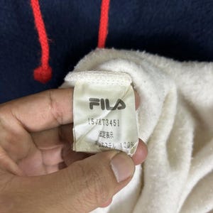 May include: Close-up of a navy blue hoodie with red drawstrings. The interior is white, and a tag with the FILA logo and text is visible. The tag also includes the text "15JXT3451", "法定表示", and "ポリエステル 100%"