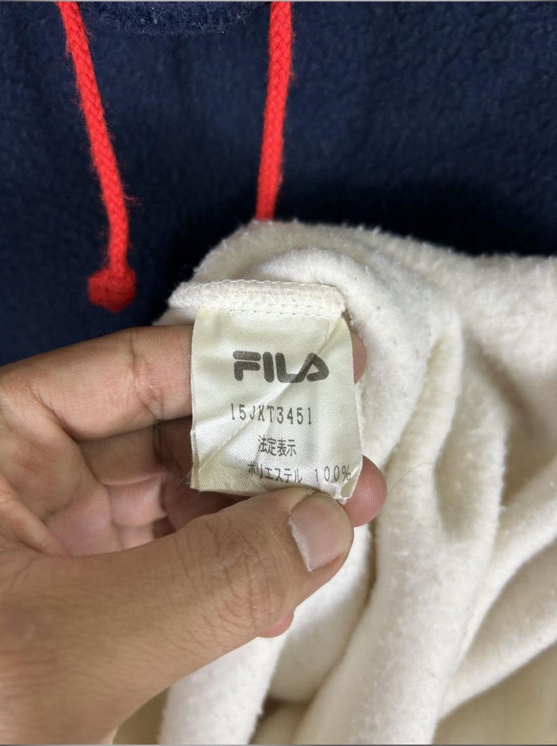 May include: Close-up of a navy blue hoodie with red drawstrings. The interior is white, and a tag with the FILA logo and text is visible. The tag also includes the text "15JXT3451", "法定表示", and "ポリエステル 100%"