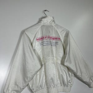 May include: White jacket with a high collar and a zipper closure. The back of the jacket features the brand name "courr&egrave;ges" in pink lettering. The jacket has a ribbed hem and cuffs. The material has a subtle sheen.