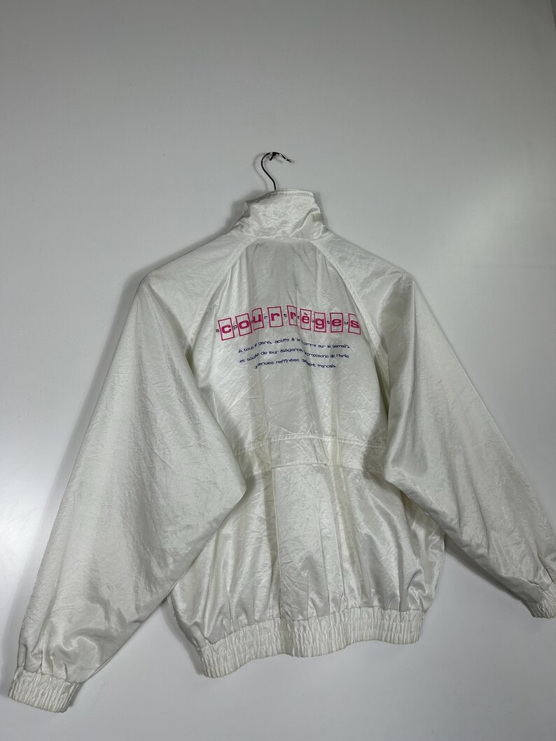 May include: White jacket with a high collar and a zipper closure. The back of the jacket features the brand name "courr&egrave;ges" in pink lettering. The jacket has a ribbed hem and cuffs. The material has a subtle sheen.