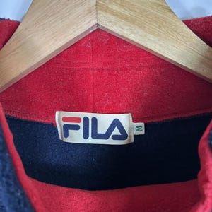May include: A close-up of a vintage Fila fleece pullover. The collar is red, with a navy blue section and a red interior. The Fila logo is visible on a tag. The garment is hanging on a wooden hanger.