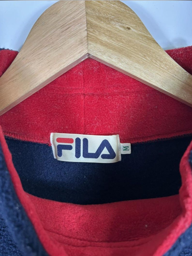 May include: A close-up of a vintage Fila fleece pullover. The collar is red, with a navy blue section and a red interior. The Fila logo is visible on a tag. The garment is hanging on a wooden hanger.
