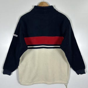 May include: A navy blue, red, and cream-colored fleece pullover. The top portion is navy blue, with a red stripe and white stripe across the chest. The bottom half is cream-colored. The Fila logo is on the left sleeve.