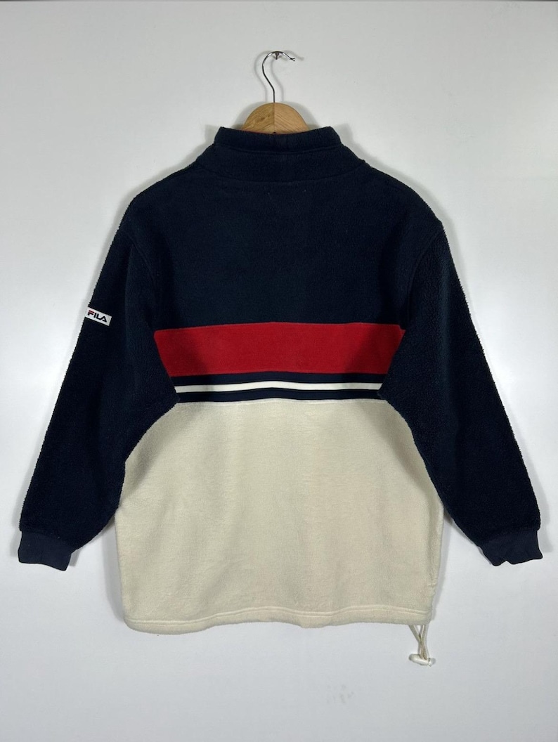 May include: A navy blue, red, and cream-colored fleece pullover. The top portion is navy blue, with a red stripe and white stripe across the chest. The bottom half is cream-colored. The Fila logo is on the left sleeve.