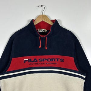 May include: A navy blue, red, and cream-colored fleece pullover. The top section is navy blue, with a red band across the chest that reads "FILA SPORTS AUTHENTIC APPAREL" in navy blue. The bottom section is cream-colored.