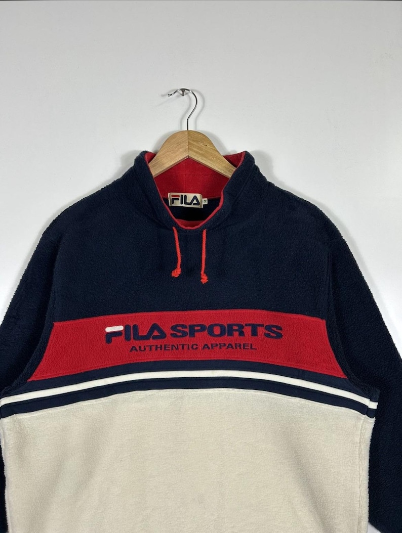 May include: A navy blue, red, and cream-colored fleece pullover. The top section is navy blue, with a red band across the chest that reads "FILA SPORTS AUTHENTIC APPAREL" in navy blue. The bottom section is cream-colored.