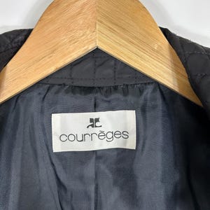 May include: Close-up of a black quilted jacket interior with a white label that reads "courr&egrave;ges" in a black font. The jacket is hanging on a wooden hanger. The interior fabric is a dark gray color.