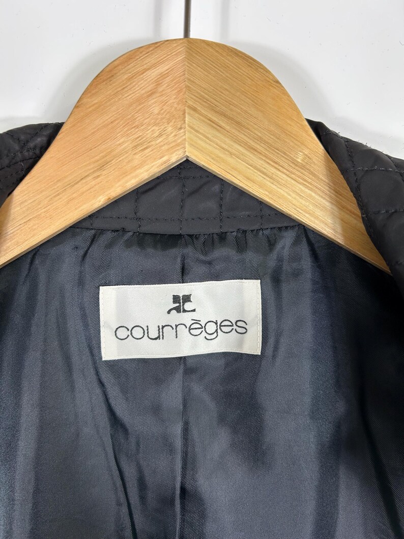 May include: Close-up of a black quilted jacket interior with a white label that reads "courr&egrave;ges" in a black font. The jacket is hanging on a wooden hanger. The interior fabric is a dark gray color.