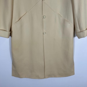 May include: A cream-coloured coat featuring a smooth texture, long sleeves with folded cuffs, and a front closure with three round, pearl-like buttons. The garment includes decorative stitched seams that radiate from the top button.