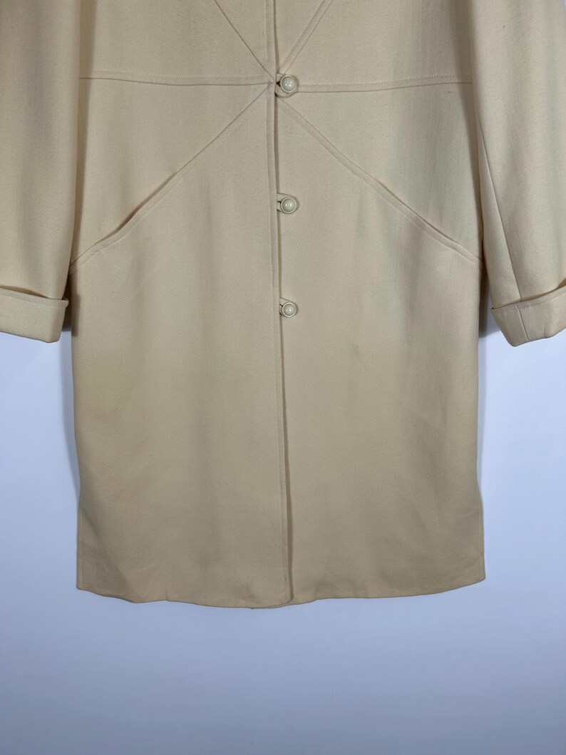 May include: A cream-coloured coat featuring a smooth texture, long sleeves with folded cuffs, and a front closure with three round, pearl-like buttons. The garment includes decorative stitched seams that radiate from the top button.